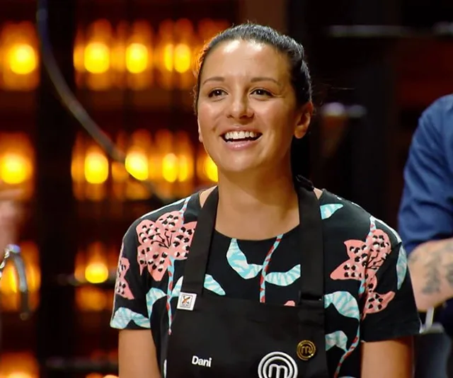 Dani from MasterChef 2020 smiling, wearing a floral shirt and a black apron, standing in a warmly lit kitchen.