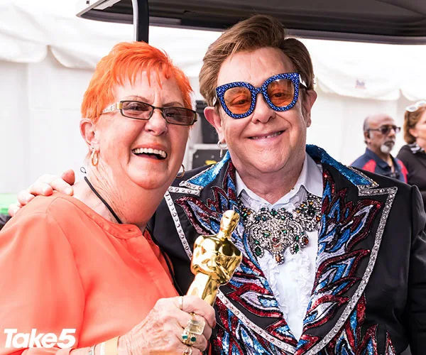 Two people smiling, one holding a gold statuette, with colorful attire and glasses, taken at an outdoor event.