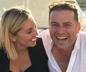 A couple smiling together, the woman looking at the man who is laughing, both in a sunny outdoor setting.