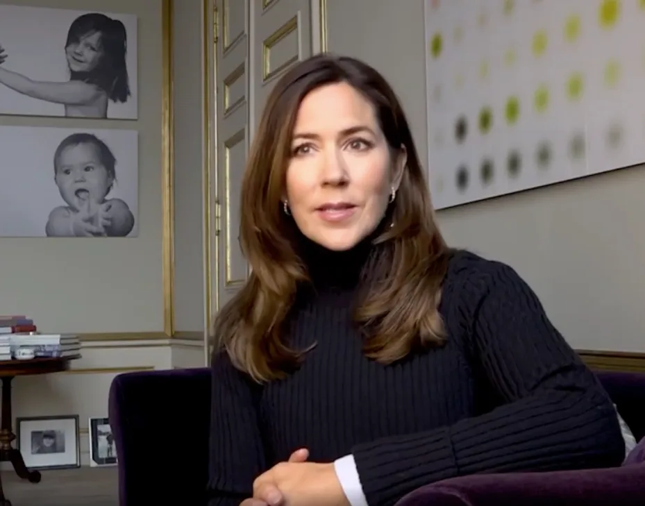 Princess Mary sitting on a chair indoors, talking, with framed child photos on the wall behind her.