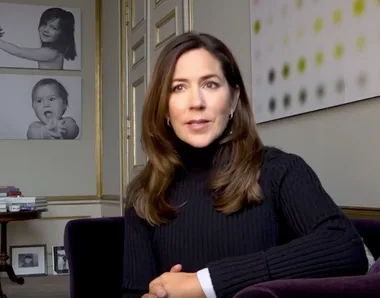 Princess Mary sitting on a chair indoors, talking, with framed child photos on the wall behind her.