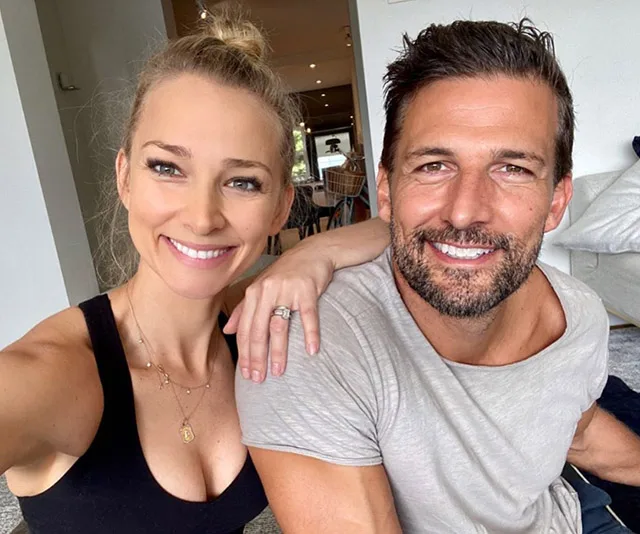 A smiling couple in casual clothes sitting together, inside a modern home, with the woman resting her arm on the man's shoulder.