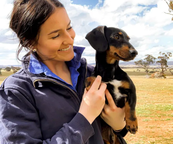 A woman smiling at a dachshund puppy outdoors, with a rural landscape in the background.