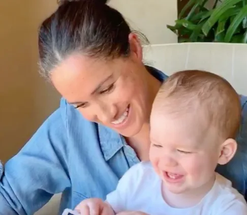 Mother in a denim shirt smiling at her baby, who is also smiling, seated together in a bright room with plants.