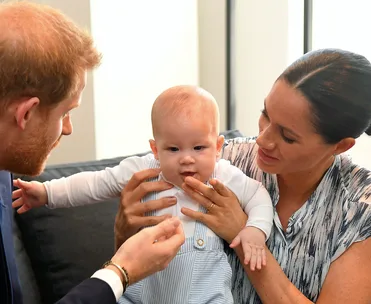 A couple lovingly holds their baby dressed in blue overalls, while smiling and sitting on a couch.