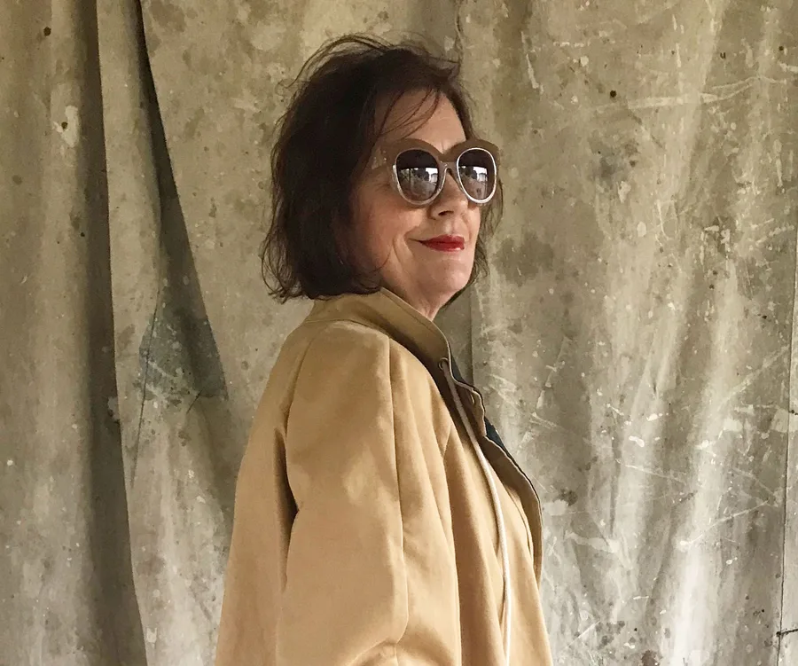 A woman wearing sunglasses and a beige coat stands in front of a textured backdrop, looking to the side.