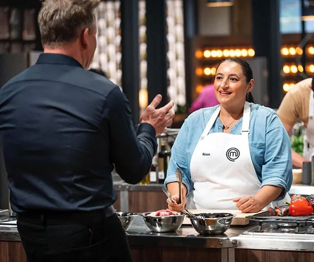 Contestant Rose, in a MasterChef apron, interacts with a chef in a kitchen during MasterChef 2020.