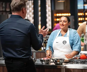 Contestant Rose, in a MasterChef apron, interacts with a chef in a kitchen during MasterChef 2020.