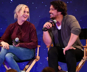 Two people smiling at each other on stage, both holding microphones, seated on director's chairs against a starry backdrop.