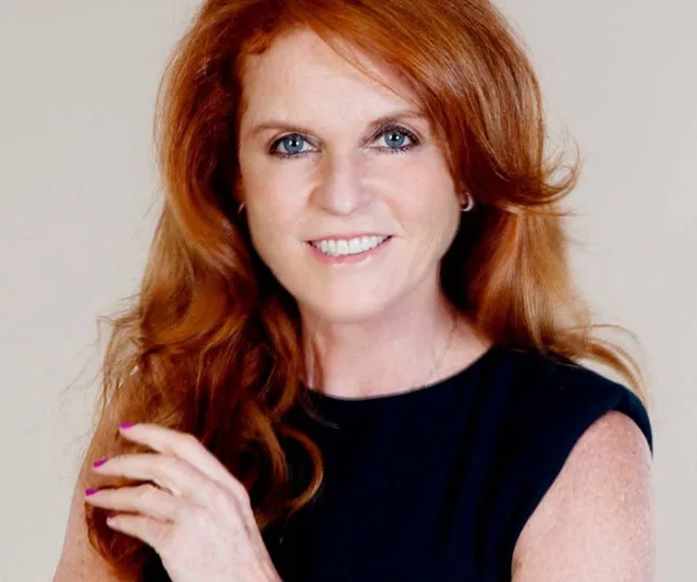A woman with long red hair and a black top smiles at the camera against a neutral background.