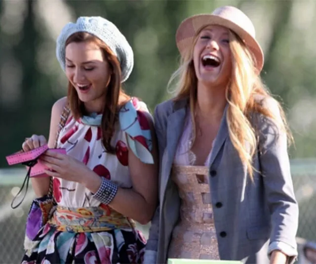Two women from Gossip Girl smiling and laughing outdoors, one holding an item, both dressed fashionably.