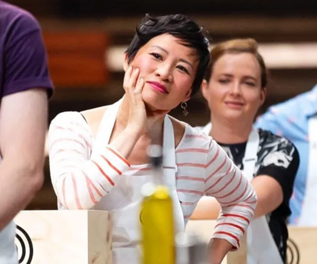 Two contestants in aprons during a MasterChef challenge, one resting her face on her hand, smiling thoughtfully.