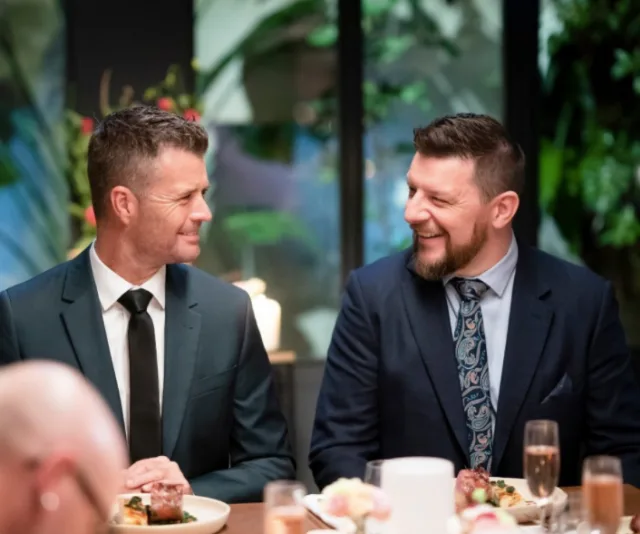 Two men in suits, smiling at each other, seated at a dinner table with plates of food and glasses in front of them.