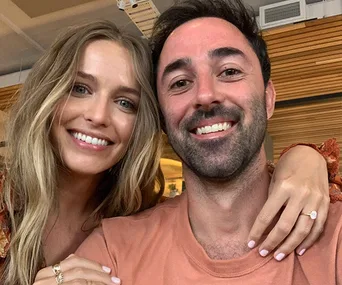 A smiling couple poses for a selfie indoors, with the woman showing an engagement ring.