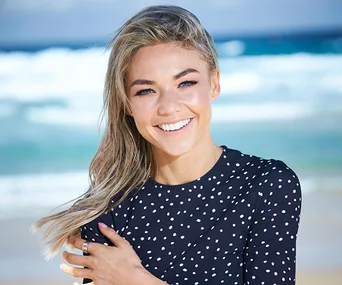 Portrait of a woman with long hair, smiling on a beach, wearing a black polka dot top with ocean waves in the background.