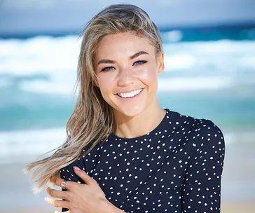 Portrait of a woman with long hair, smiling on a beach, wearing a black polka dot top with ocean waves in the background.