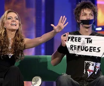 A person with taped mouth holds a "Free the Refugees" sign, next to another person gesturing on a TV set.