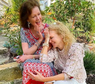 Two women sitting outside smiling, one wearing a red-patterned dress and the other a floral blouse, sharing a tender moment.