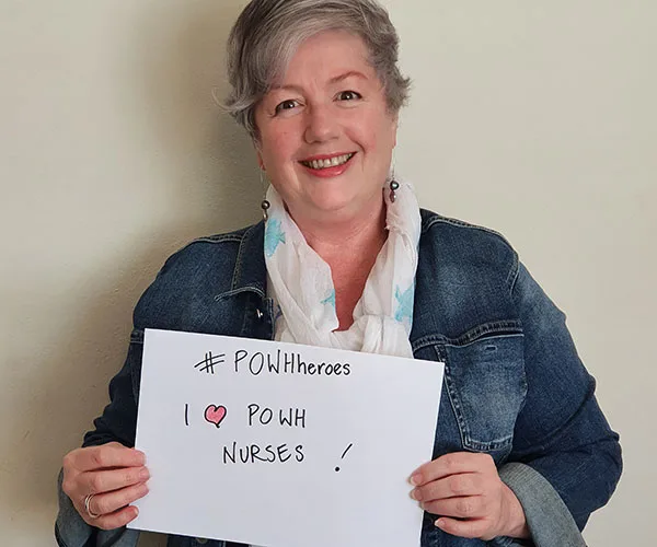 A person holding a sign with the text "#POWHeroes I ♥ POWH Nurses!" while smiling at the camera.
