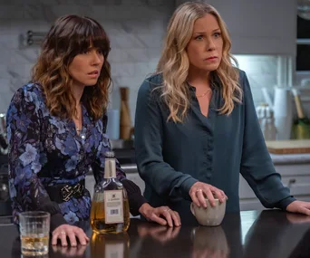 Two women, one in a floral dress and the other in a blue blouse, stand in a kitchen with drinks on the counter.