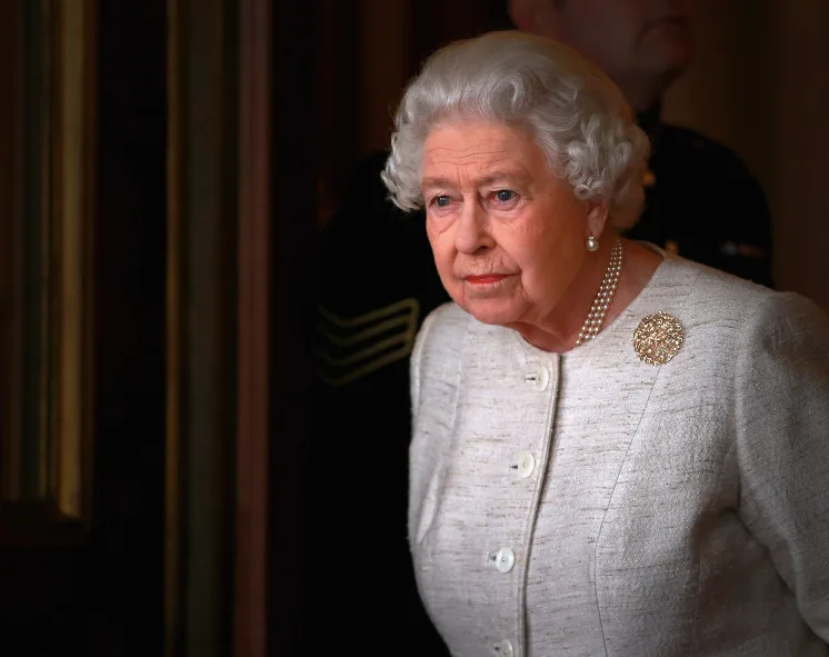 Queen Elizabeth II in a white suit with pearls and brooch, looking serious, standing near a doorway.