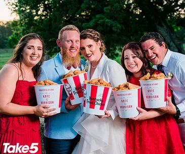 Five people at a wedding holding KFC buckets labeled "Congratulations Kate & Harrison," smiling outdoors.