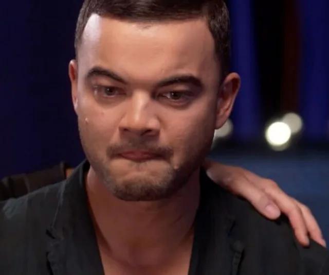 A man with short hair, emotional, with tears, is consoled by a hand on his shoulder against a blurred background.