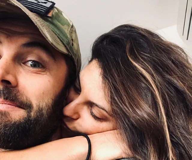 A couple in a playful embrace; the man wears a camo cap, while the woman snuggles close, eyes closed.