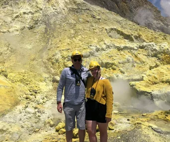 Couple with helmets posing on a rocky, sulfur-covered landscape with steam rising, likely White Island.
