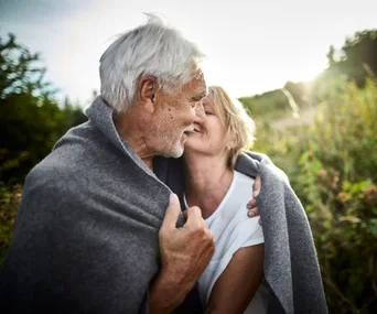 Elderly couple wrapped in a blanket, embracing and smiling outdoors with sunlight in the background.
