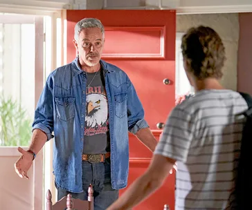 Man in denim shirt stands by a red door, talking to a person in a striped shirt.