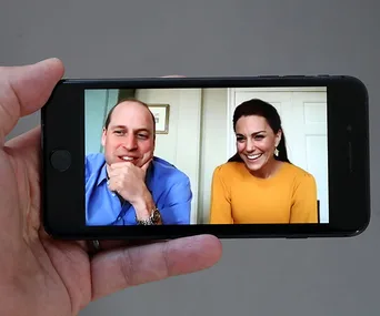 A hand holds a smartphone showing a video call between a man in a blue shirt and a woman in a yellow top, both smiling.