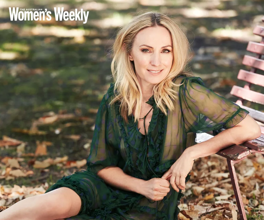 A woman with blonde hair sits on a pink bench outdoors, wearing a sheer green dress, with leaves scattered on the ground.