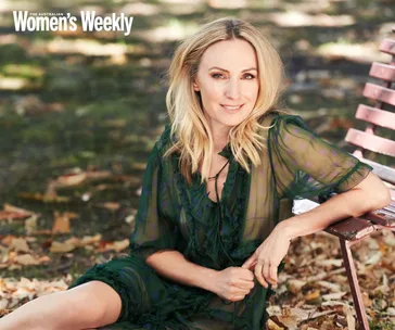 A woman with blonde hair sits on a pink bench outdoors, wearing a sheer green dress, with leaves scattered on the ground.