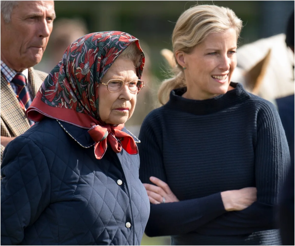 Two women outdoors, one in a floral headscarf and glasses, the other in a dark sweater, with a man in the background.