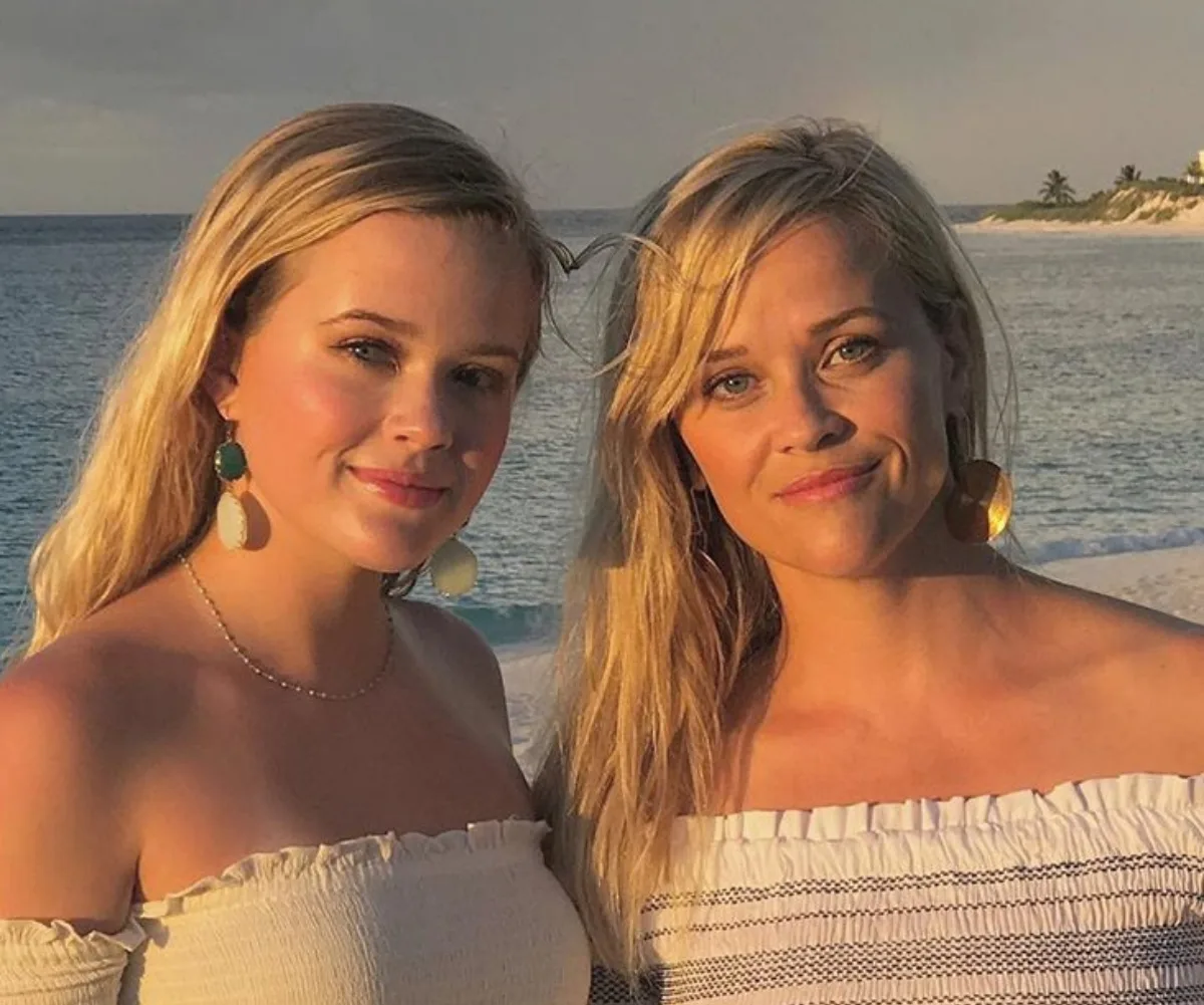 Two women with long blonde hair, wearing off-shoulder tops, stand on a beach at sunset.