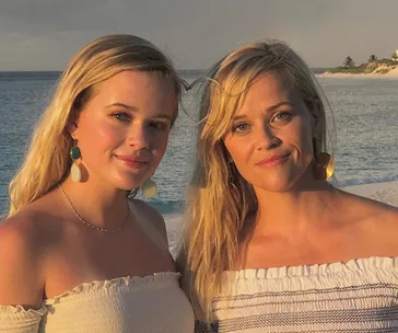 Two women with long blonde hair, wearing off-shoulder tops, stand on a beach at sunset.