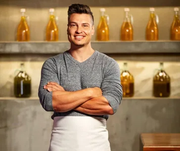 A man in a gray sweater and white apron, arms crossed, standing in a kitchen with shelves of bottled liquids in the background.