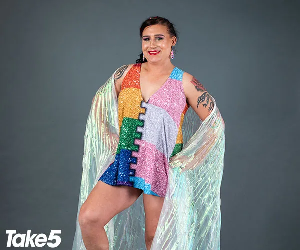 Transgender individual in a colorful patchwork dress with tattoos and iridescent cape, smiling confidently at the camera.