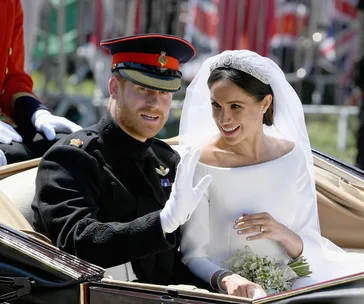 Prince in uniform and bride in white dress with bouquet, smiling in a carriage during wedding procession.