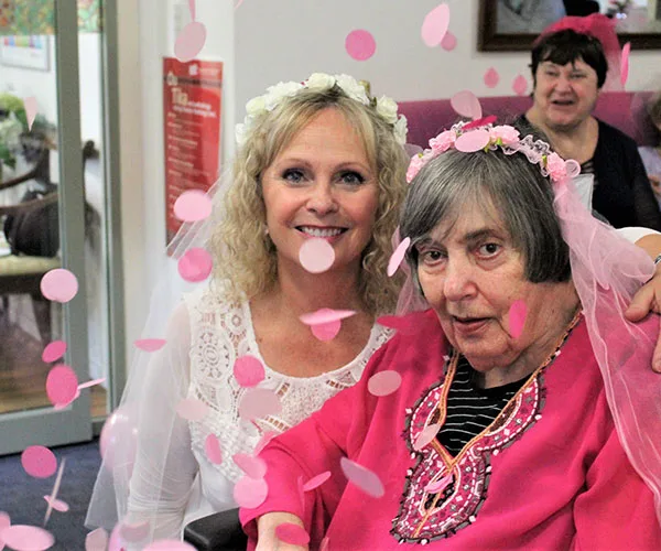 Two women at a festive gathering with pink confetti and flower crowns, one in a wheelchair, smiling at the camera.
