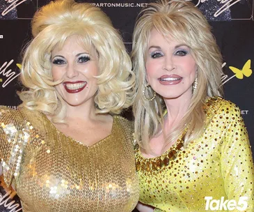 Two women in shiny dresses with blonde hair at an event, smiling and posing together.