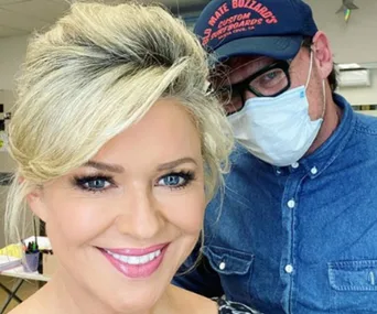 Blonde woman smiling, posing with a man in a denim shirt and mask inside a bright room.