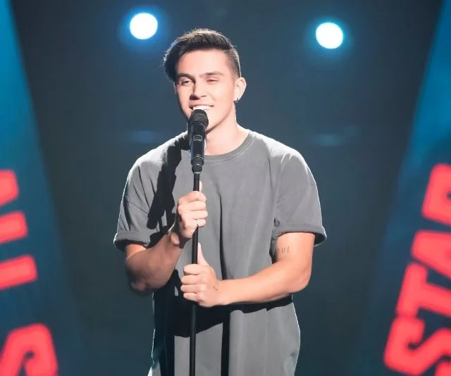 Singing contestant on stage under spotlights with microphone in hand.