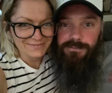 Two people, a woman with glasses and a man with a beard, smiling closely together for a selfie indoors.