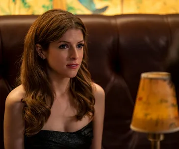 A woman with long hair sits on a leather couch, looking thoughtful, with a yellow lamp in the foreground.