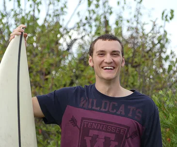 Man smiling, holding a surfboard, wearing a "Wildcats Tennessee" shirt, standing outdoors with greenery in the background.