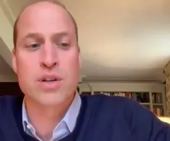 A person speaking on a video call, seated in a room with bookshelves.