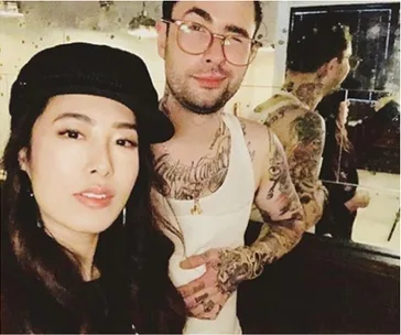 A woman in a black hat takes a selfie with a tattooed man in glasses and a tank top standing beside her.