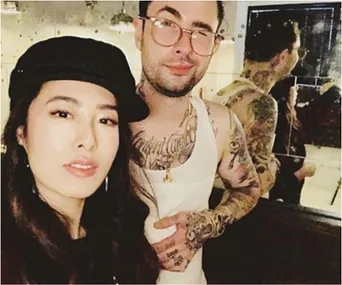 A woman in a black hat takes a selfie with a tattooed man in glasses and a tank top standing beside her.